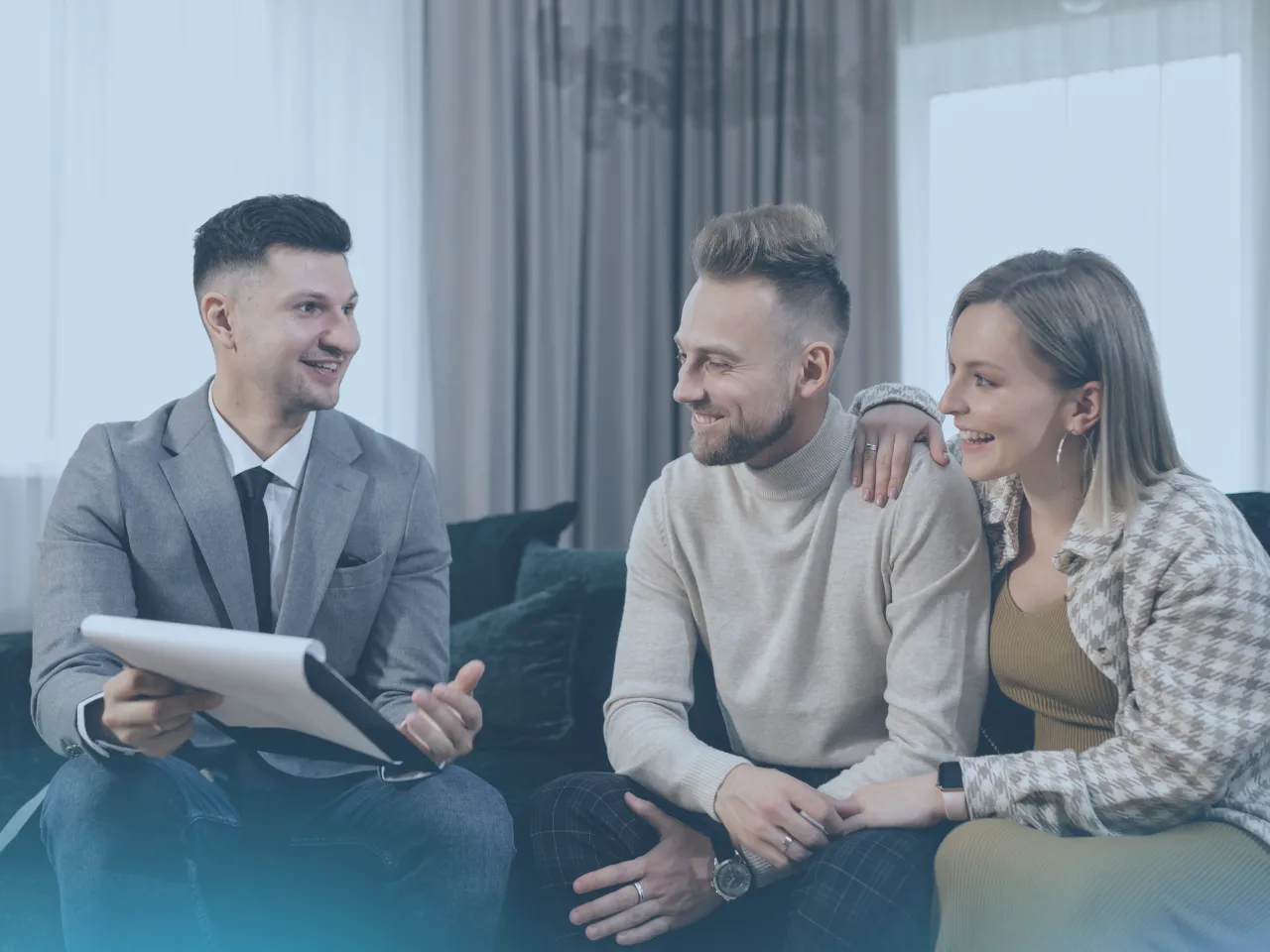 A male real estate agent in a grey suit and black tie sits across from a smiling couple on a couch. The agent is holding a folder and gesturing with his left hand, talking to the couple, who are looking at him attentively.