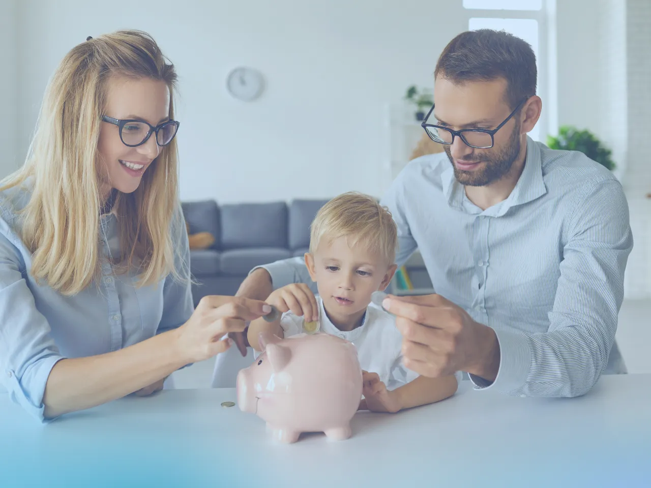 A family of three, a mother, father, and young son, are sitting at a table. The mother and son are placing coins into a pink piggy bank, while the father also holds a coin, looking on. All three are smiling.
