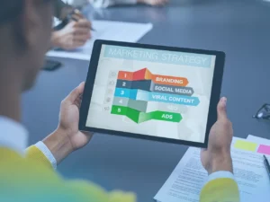 A person is holding a tablet that displays a "Marketing Strategy" flowchart. The flowchart shows a list of five key components, each with an icon and a colored block: 1. Branding, 2. Social Media, 3. Viral Content, 4. SEO, and 5. Ads. Other documents are visible on the table.