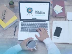 An overhead view of a person's hands on a laptop keyboard. The laptop screen displays a web page with a bullseye target icon and the words "Target Audience." The person is holding a coffee cup with one hand. Various office supplies like books, a pen, glasses, and a smartphone are scattered on the wooden desk.