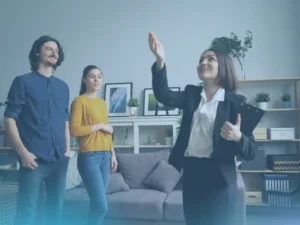 A smiling female real estate agent in a dark blazer and white shirt gestures upwards with her hand while showing a living room to a young couple. The couple, a man in a blue shirt and a woman in a yellow sweater, stand beside her and look towards where she is pointing.