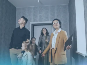 A real estate agent in a brown blazer, holding a folder, smiles as she shows a family of four a home. The family consists of a father, a mother, and two young daughters. They are all looking up and around the room with expressions of interest and excitement.